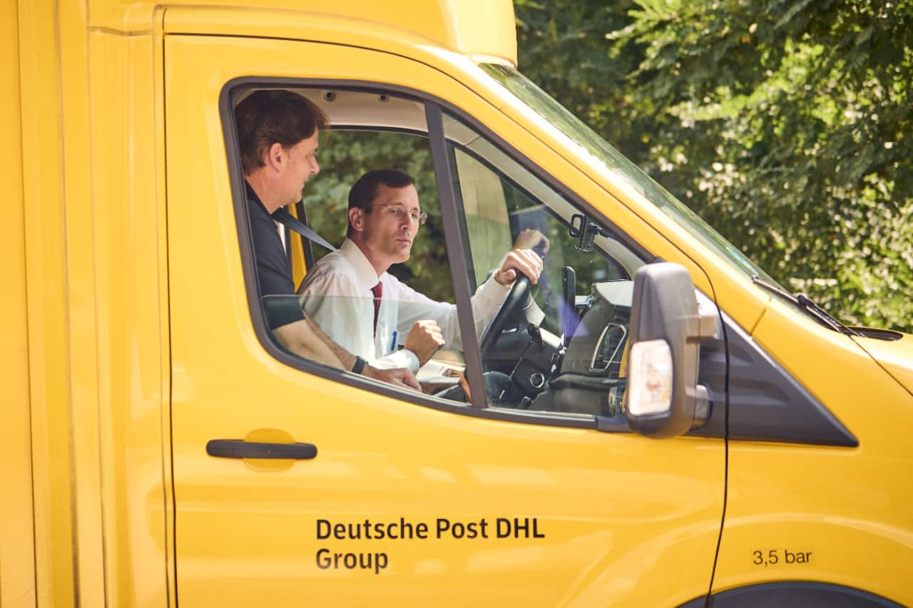 Two men sit in the front seats of a yellow DHL E-Transit.